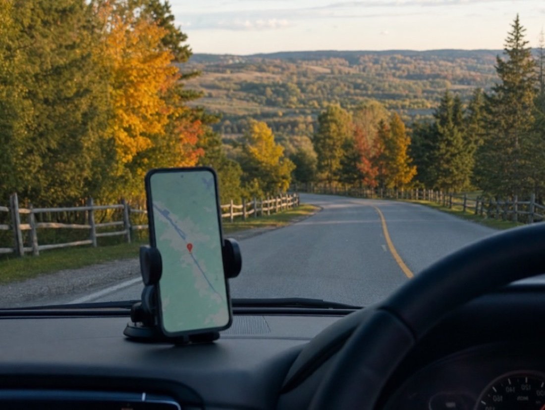 Phone in a car mount showing a red map pin, the two-lane road ahead running through autumn trees